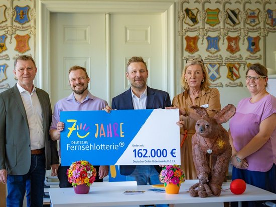 Fünf Personen halten einen großen Scheck der Deutschen Fernsehlotterie. Der Scheck über 162.000 Euro an den Deutschen Orden wird im Rahmen des 70-jährigen Jubiläums der Lotterie überreicht.