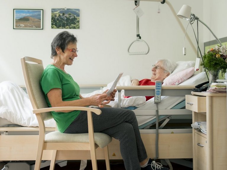 Eine Mitarbeiterin liest einer im Bett liegenden Bewohnerin etwas vor.