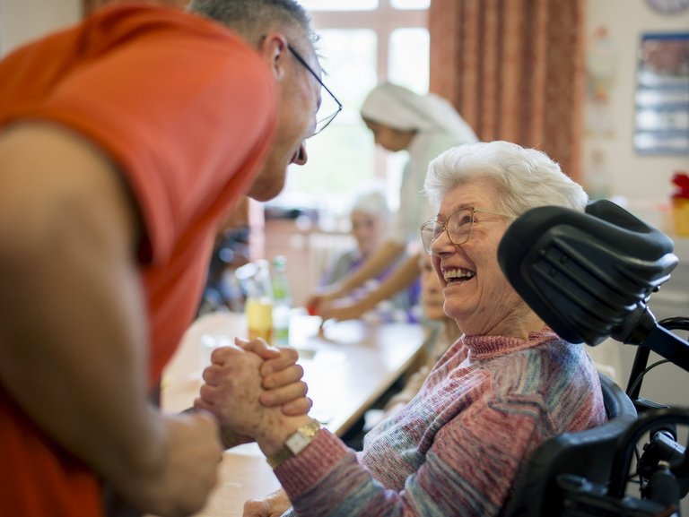 Eine Bewohnerin und ein Mitarbeiter geben sich die Hand und lächeln sich an.